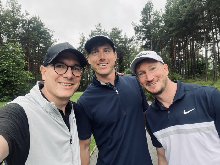 Drei Golfer stehen nach der Runde Arm in Arm auf dem Platz, dahinter Nadelwald.