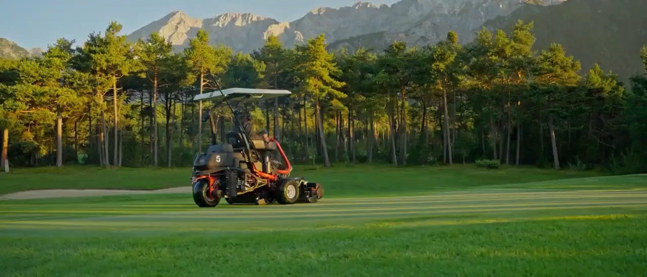 Ein Greenkeeper mäht im Morgenlicht ein Fairway, dahinter die Mieminger Kette.